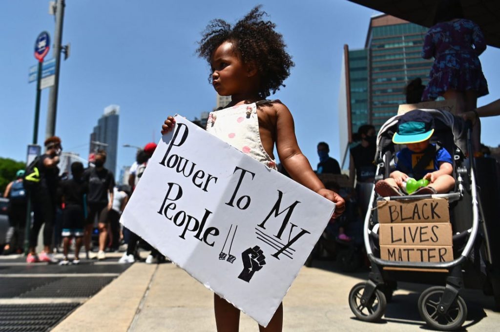 Image of a little girl at a protest holds a sign that reads "Power To My People."