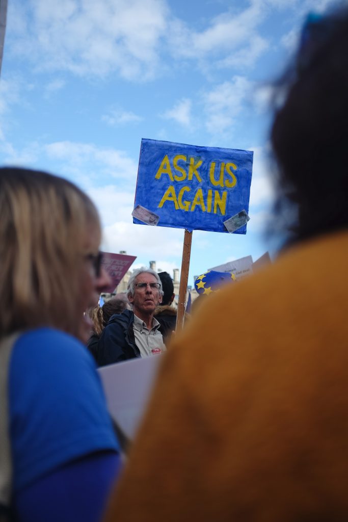 An abstract image of a man holding a poster reading 'Ask us again' in the context of Brexit