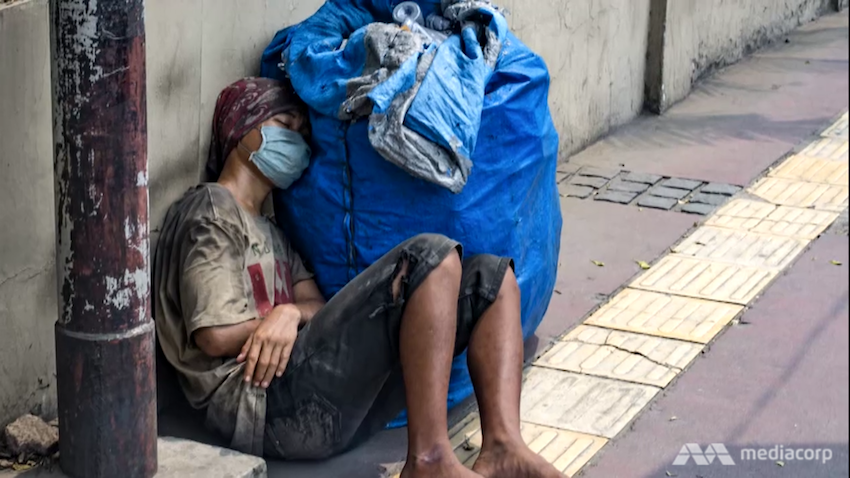 A man wearing a mask and torn clothes is resting on a sidewalk.