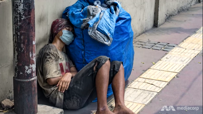 A man wearing a mask and torn clothes is resting on a sidewalk. 