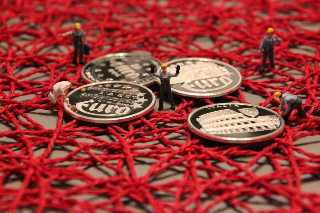 A photo of a massive tangle of red string with a few euro coins on top and tiny construction worker figurines