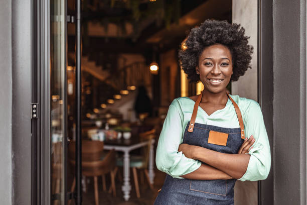 Cheerful female café owner standing at the door with her arms crossed. African woman in apron standing with her arms crossed