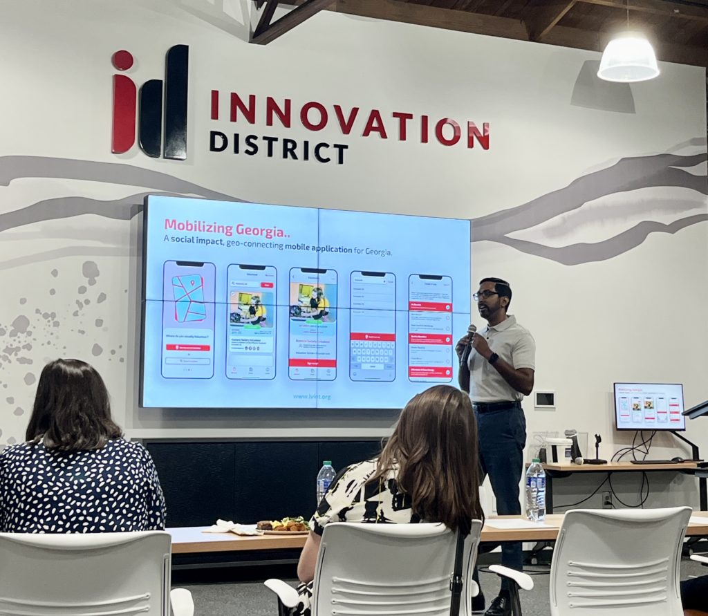 A picture of Nipuna Ambanpola talking with a microphone in his hand, with the IVolunteer International mobile app screens behind in, on a screen.